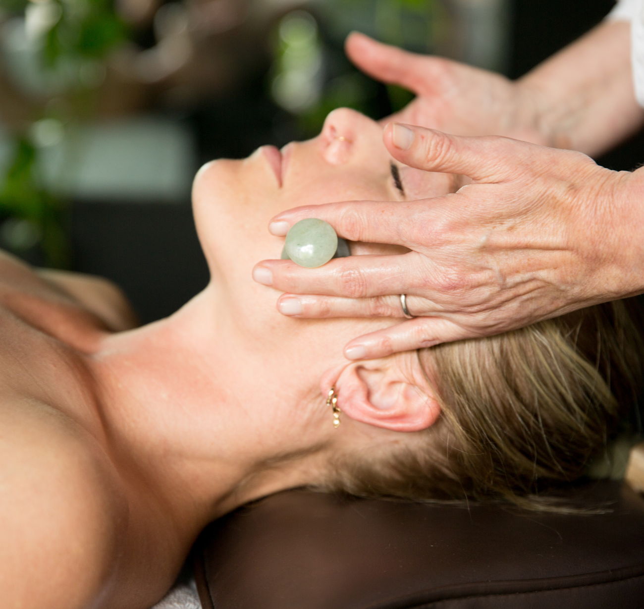 A patient resting with eyes closed while jade Gua Sha tools are worked across the face
